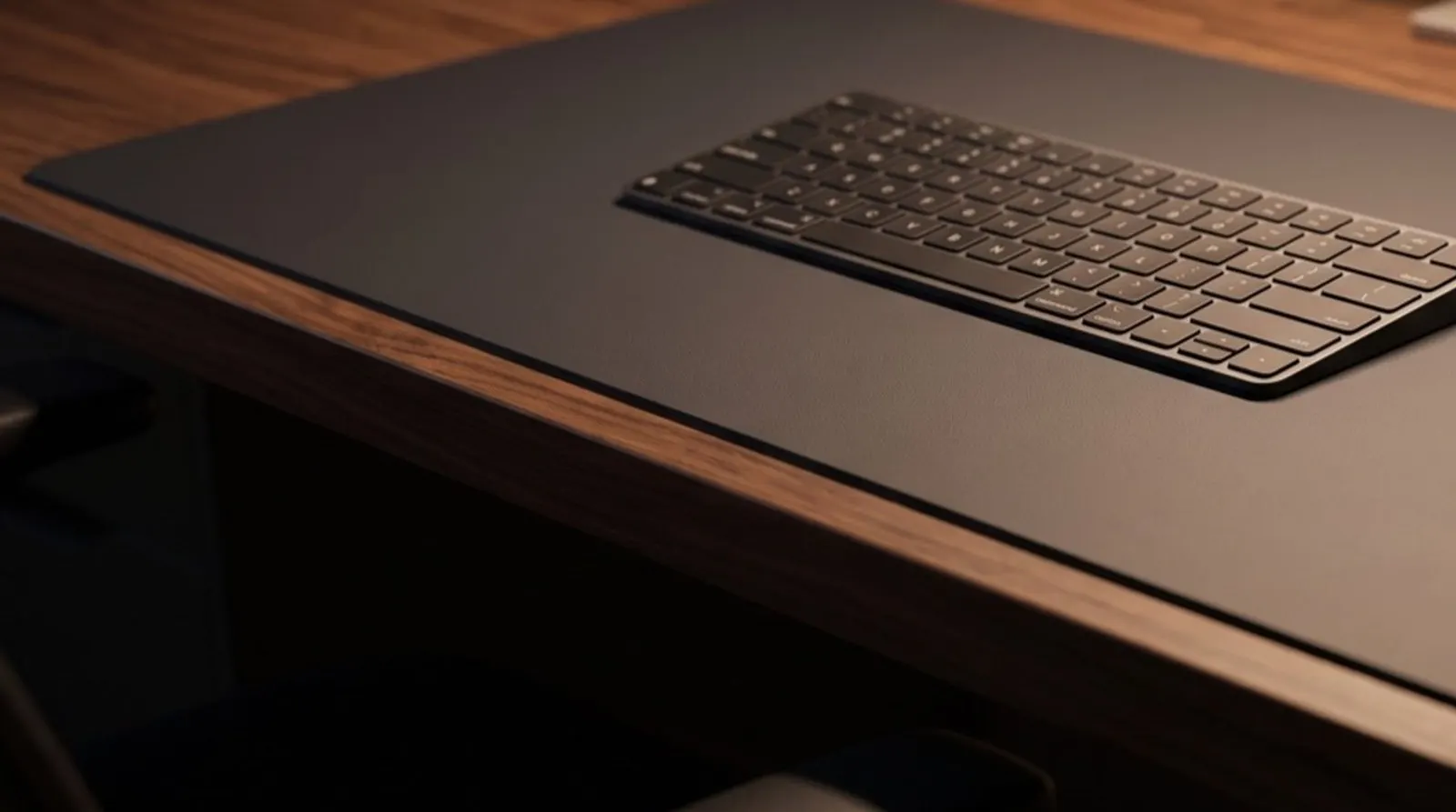 デスクマットの端が天板の縁に沿ってまっすぐ敷かれている様子のクローズアップ。マットの上にキーボードが整然と置かれ、暖色の照明が当たっている。